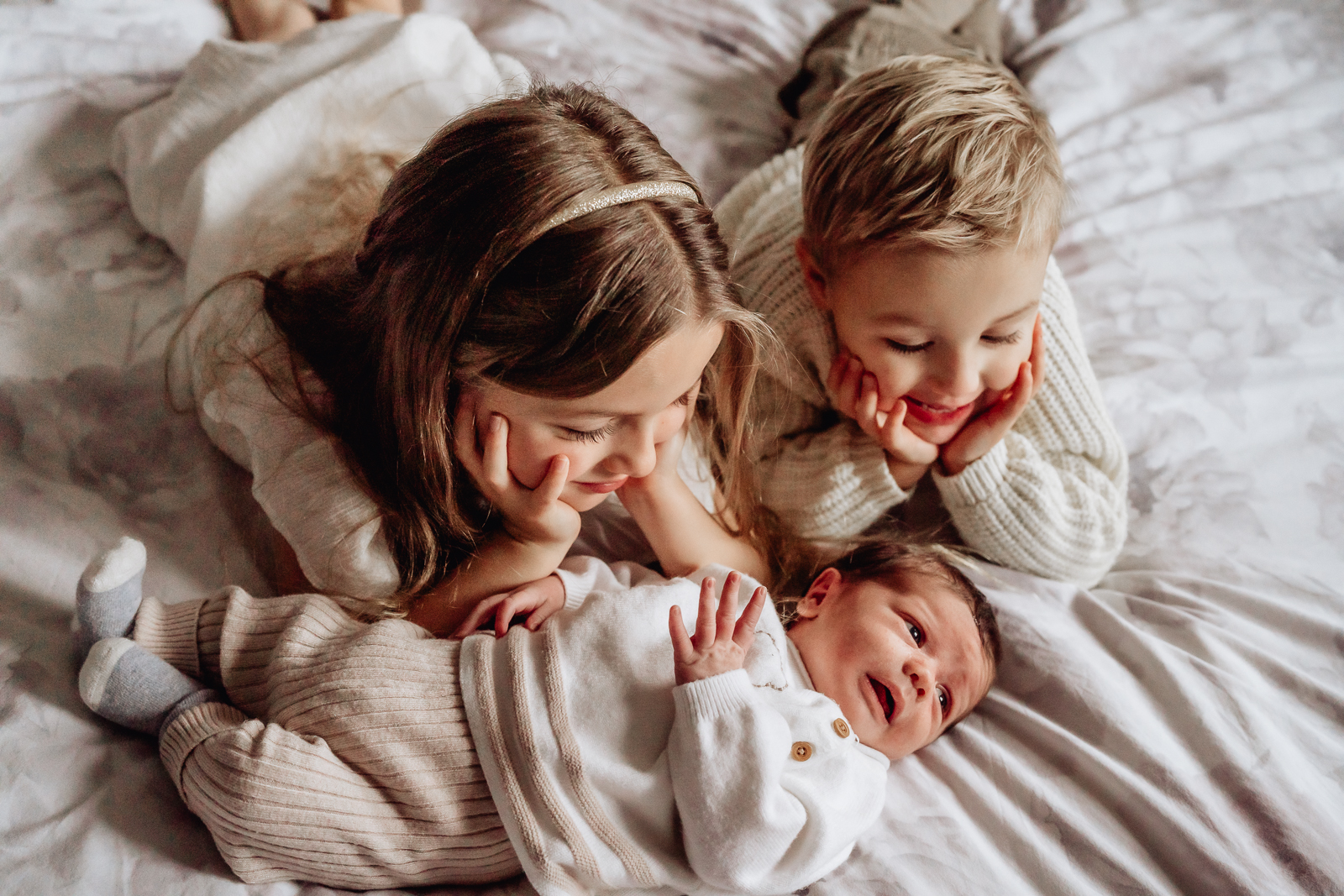 A young girl and boy are lying on their tummies on a bed next to each other, looking at a small baby who is lying in front of them on the bed