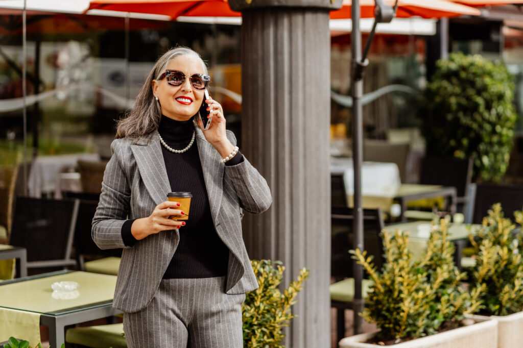 A lady in a grey suit and black top is wearing sunglasses and holding a coffee cup while she talks on the phone.